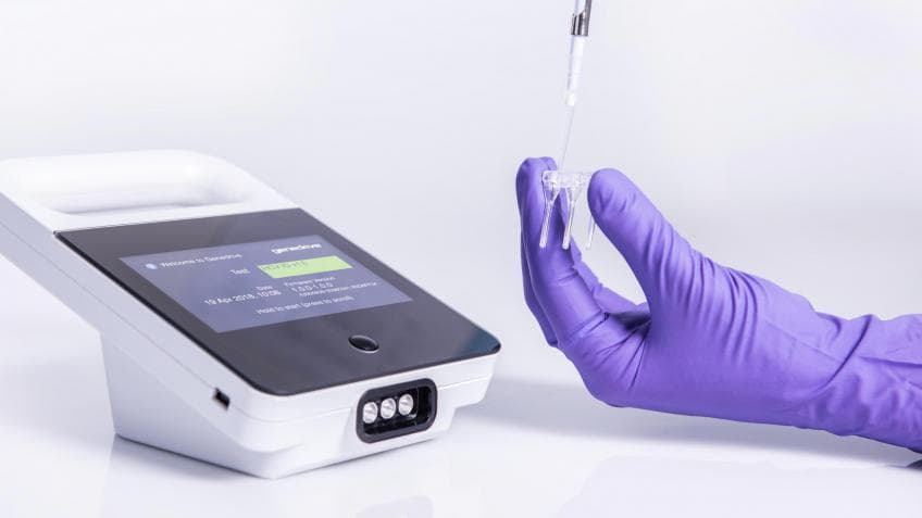A gloved hand holding a sample pipette beside a compact diagnostic device